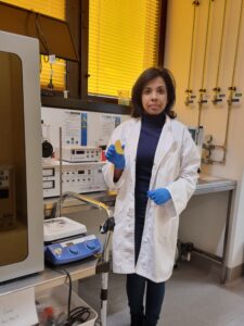 Nitu Syed holding an ammonia gas sensor that looks like a golden half moon standing in front of laboratory equipment